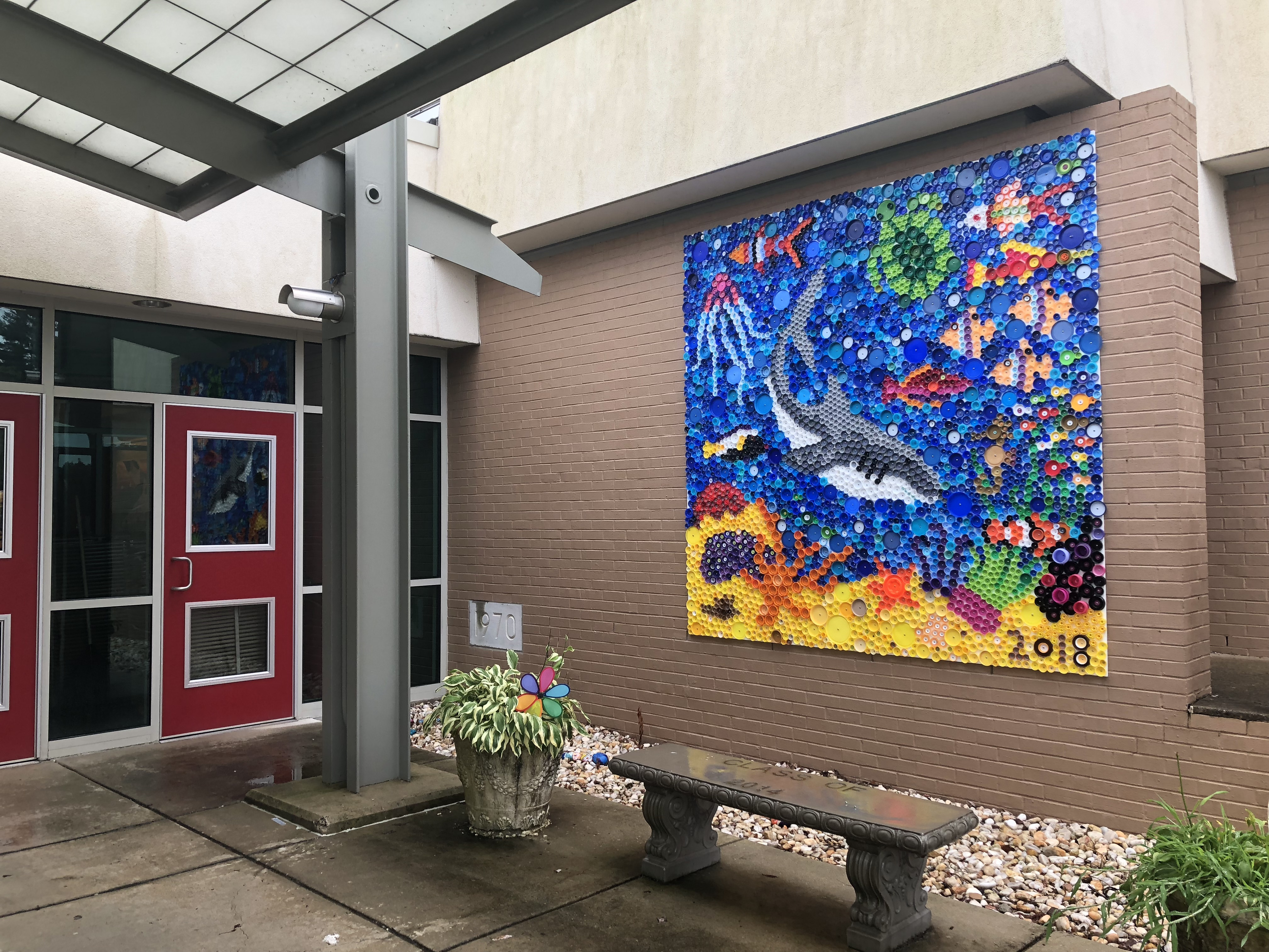 The front entrance of a tan brick elementary school is shown with two red doors, a green plant with a colorful windmill, and a concrete bench with "Class of 2014" engraved into the top. A white plaque on the building says 1970 and a large colorful mural showing a shark swimming in an underwater scene made out of bottle caps.