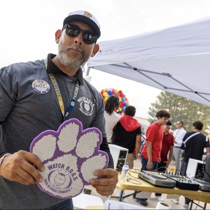 A man in the foreground holds a purple paw print while a group of people gather under a tent in the background.