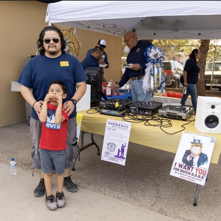 A man and a young boy stand in the foreground, while a DJ setup and people in the background are under a tent.