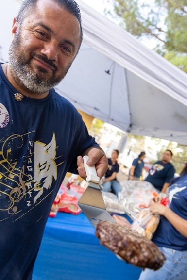 A man in the foreground holds a spatula with a burger, while people gather under a tent in the background.