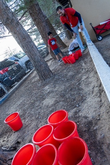 Red buckets are arranged on the ground in the foreground, while two people in red shirts play a game in the background with trees and cars visible.