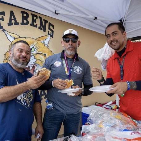 Three men stand in front of a mural of a bobcat and the word "FABENS" while holding sandwiches and plates.