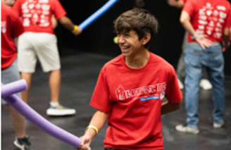 A smiling boy in a red t-shirt holds a purple foam noodle in the foreground, while other people in red shirts are blurred in the background.