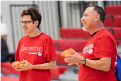 Two men in red t-shirts stand in the foreground, holding orange objects, with blurred red bleachers in the background.