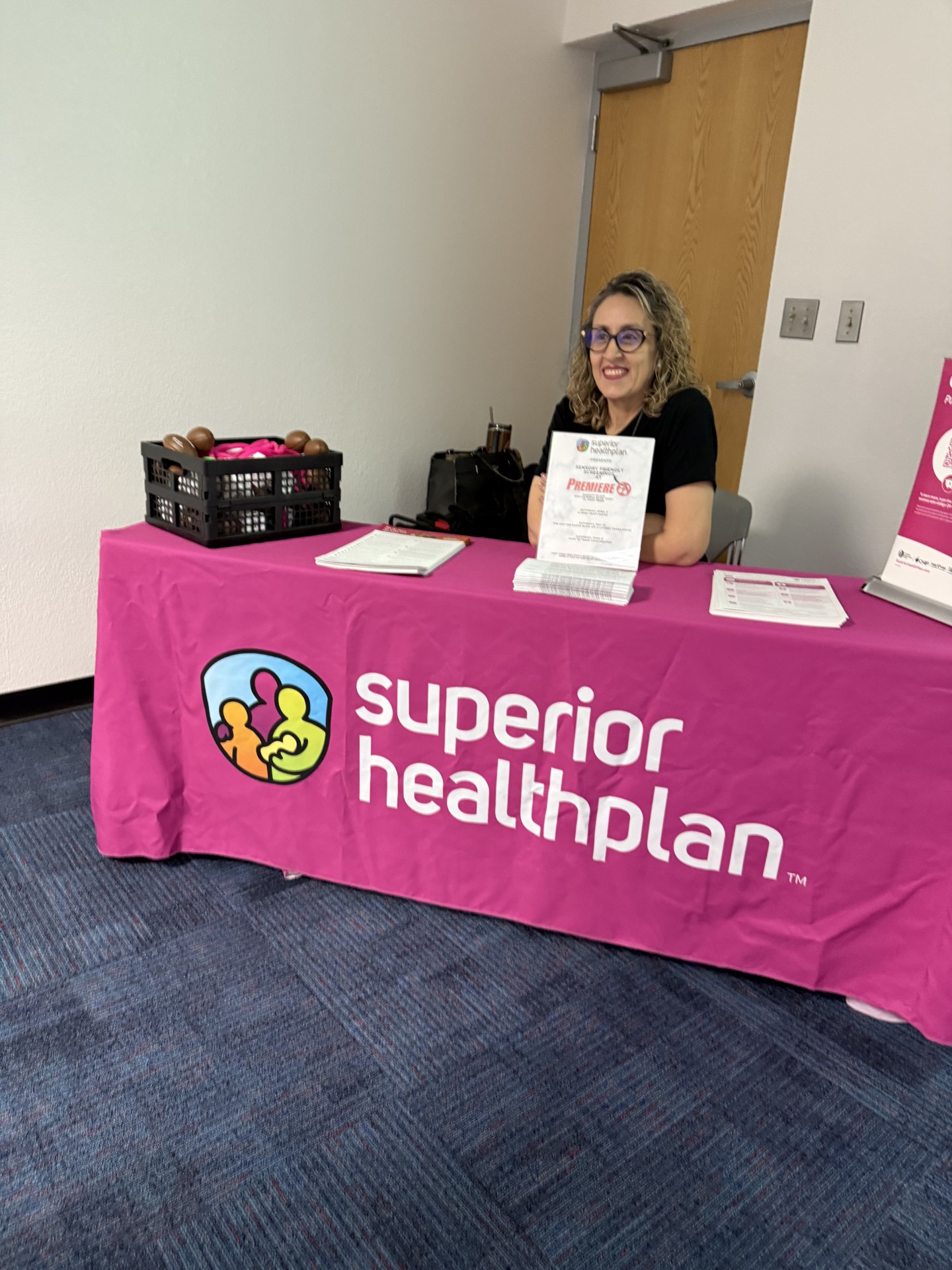 A woman with curly hair and glasses smiles from behind a pink table with the "superior healthplan" logo, while a black basket of items sits on the left side of the table in front of a plain wall.