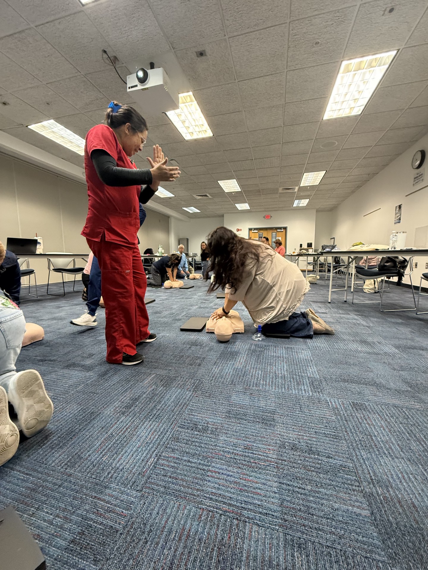 In the foreground, a woman in red scrubs demonstrates CPR on a manikin while others practice in the background of a classroom with tables and chairs.