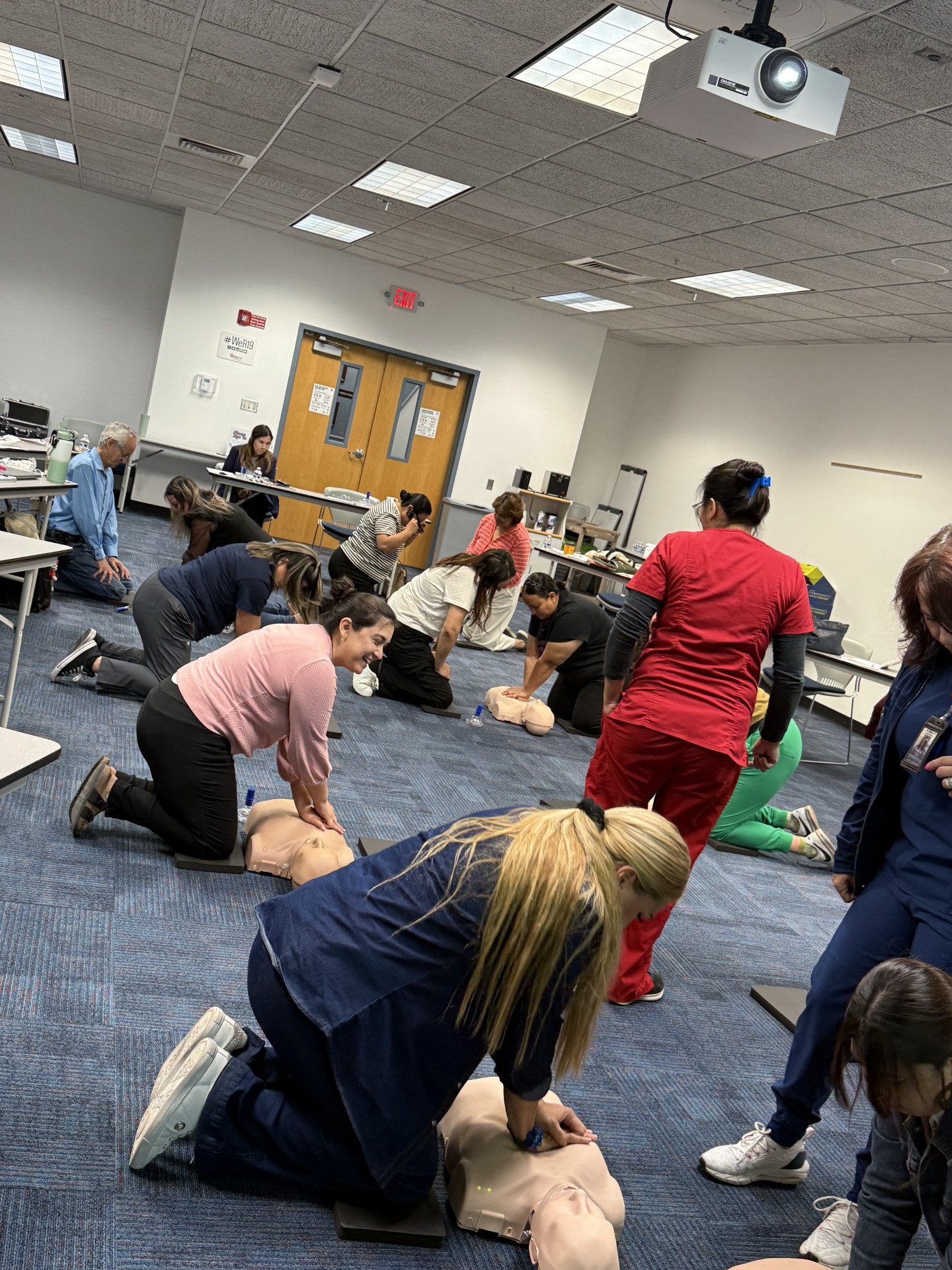 In the foreground, several people are practicing CPR on mannequins, while the background shows a classroom setting with tables, chairs, and a projector.