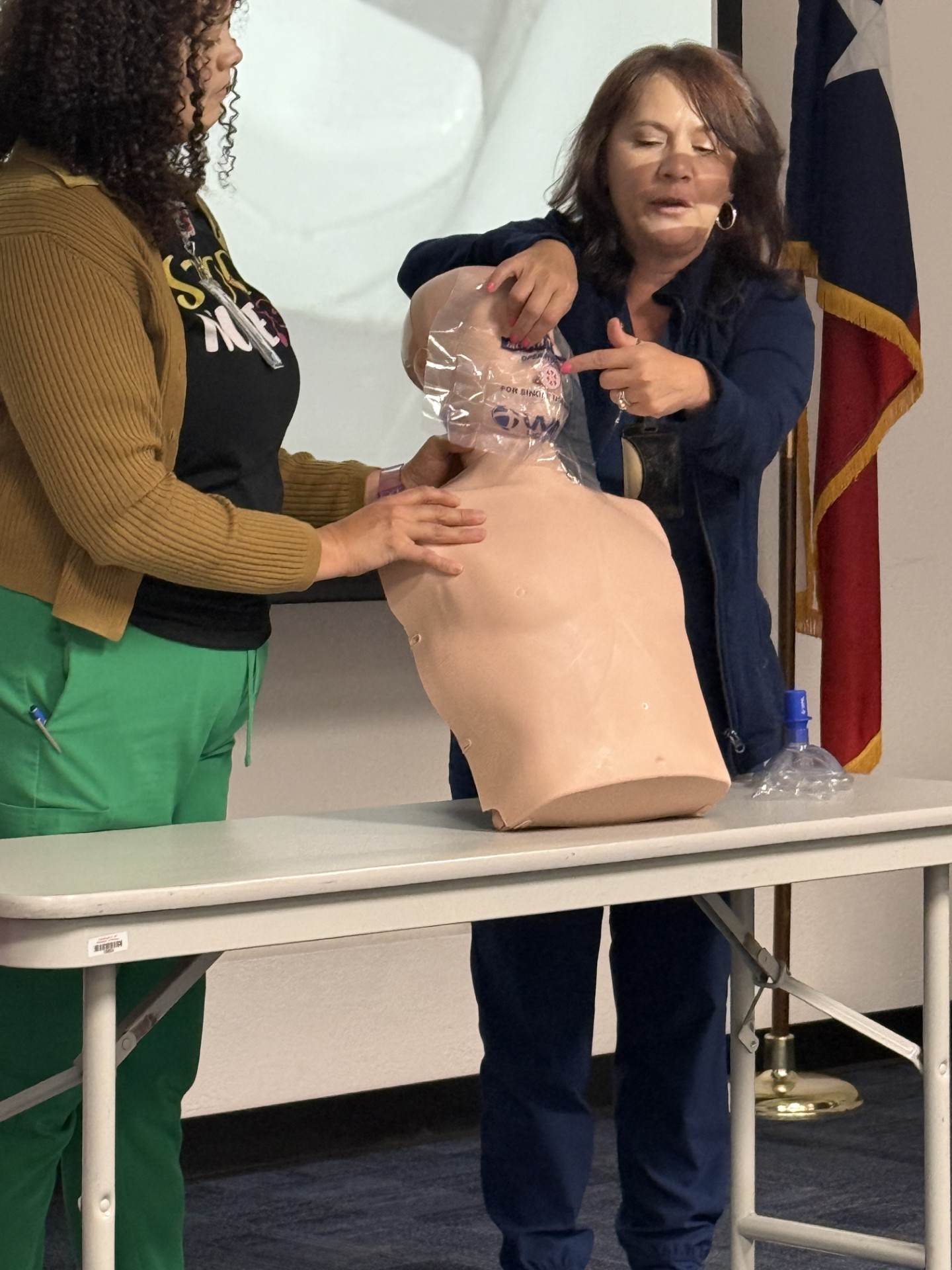 Two women are practicing CPR on a dummy in front of a projector screen and a Texas flag.