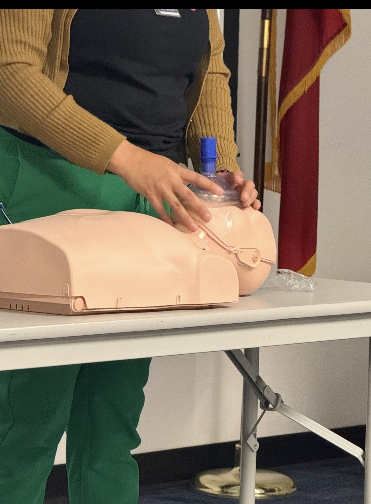 A person in green scrubs and a tan cardigan practices CPR on a manikin, with flags visible in the background.