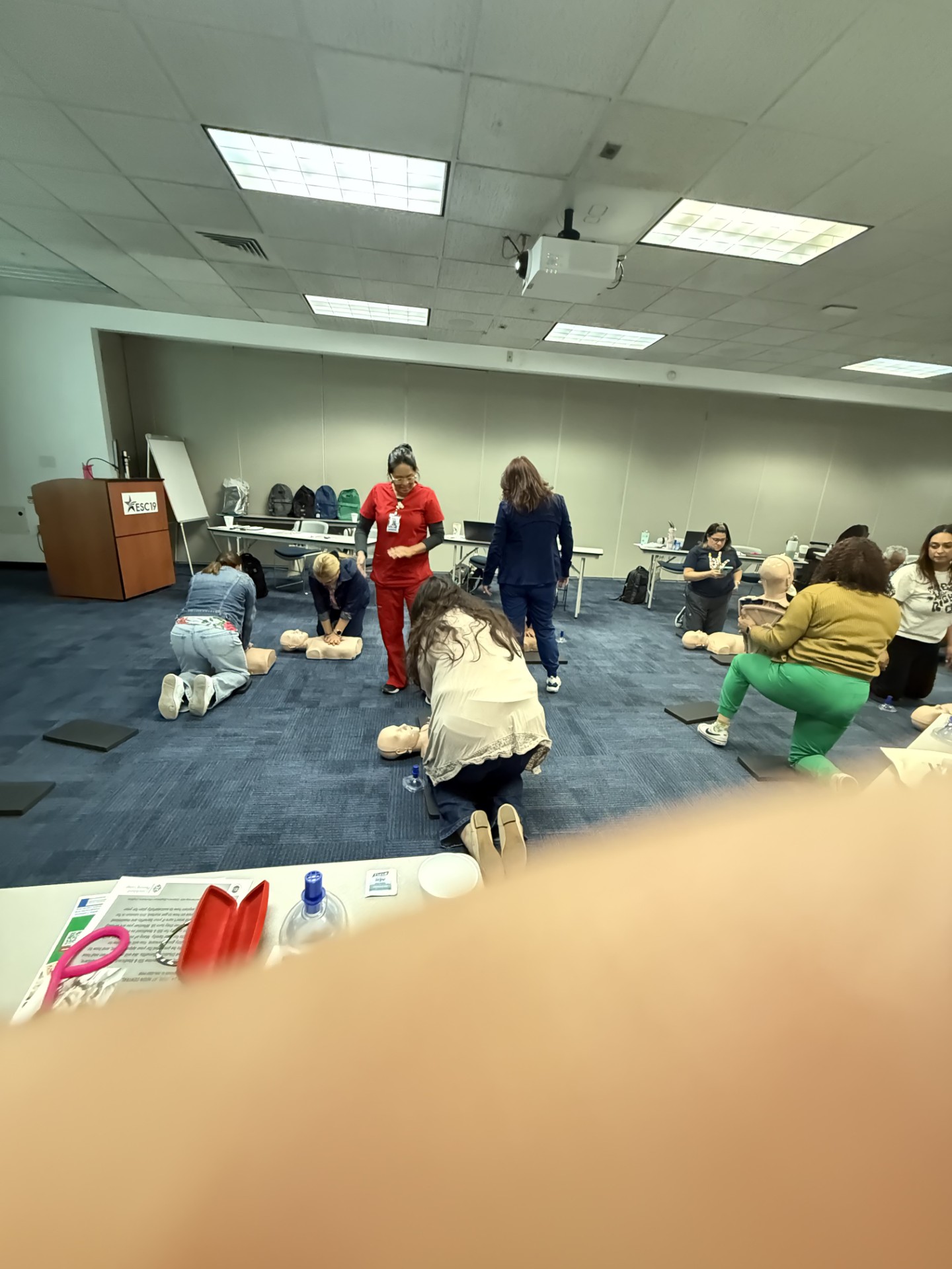 In the foreground, a blurry hand partially obscures a table with medical supplies, while in the background, a group of people practice CPR on mannequins in a classroom setting.