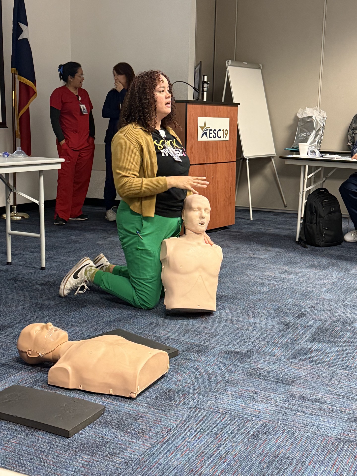 A woman in green pants demonstrates CPR on a mannequin in the foreground, while others observe in the background near a podium and whiteboard.