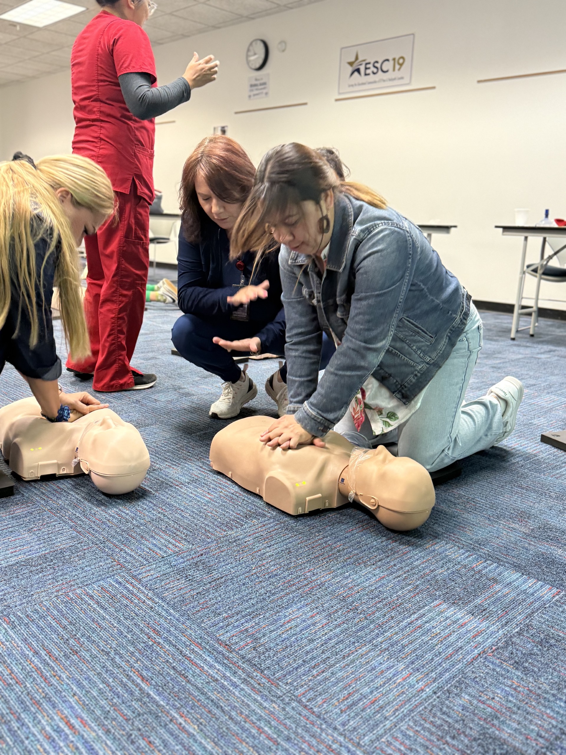 In the foreground, several people practice CPR on manikins, while in the background, a classroom setting is visible with tables and chairs.