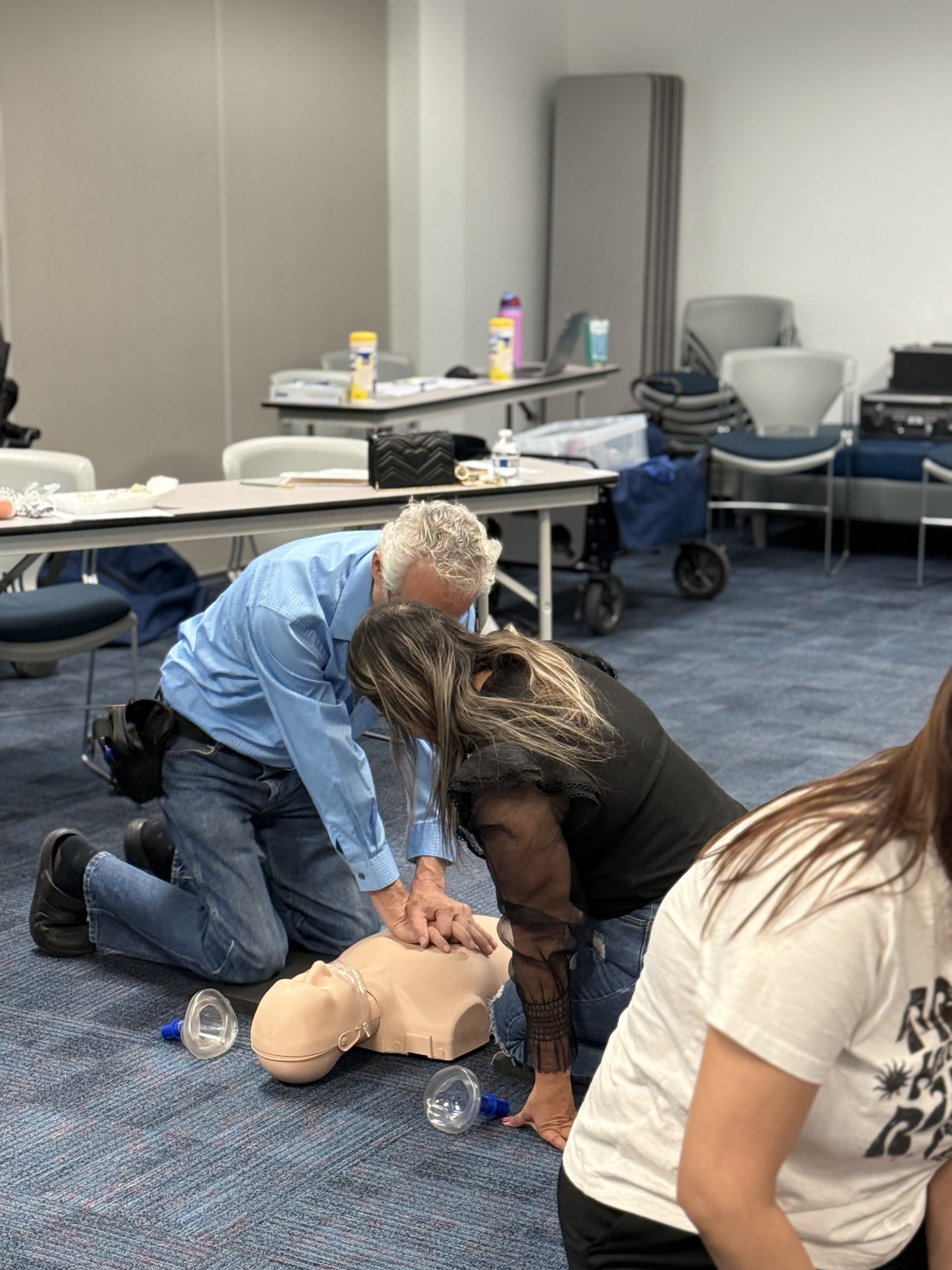 In the foreground, two people practice CPR on a manikin, while the background shows a classroom setting with tables and chairs.
