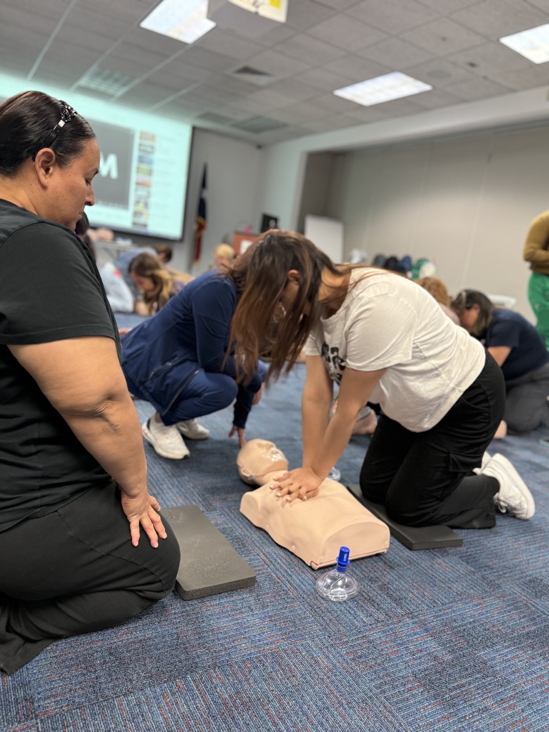In the foreground, several people practice CPR on a manikin, while in the background, a projector screen displays information.