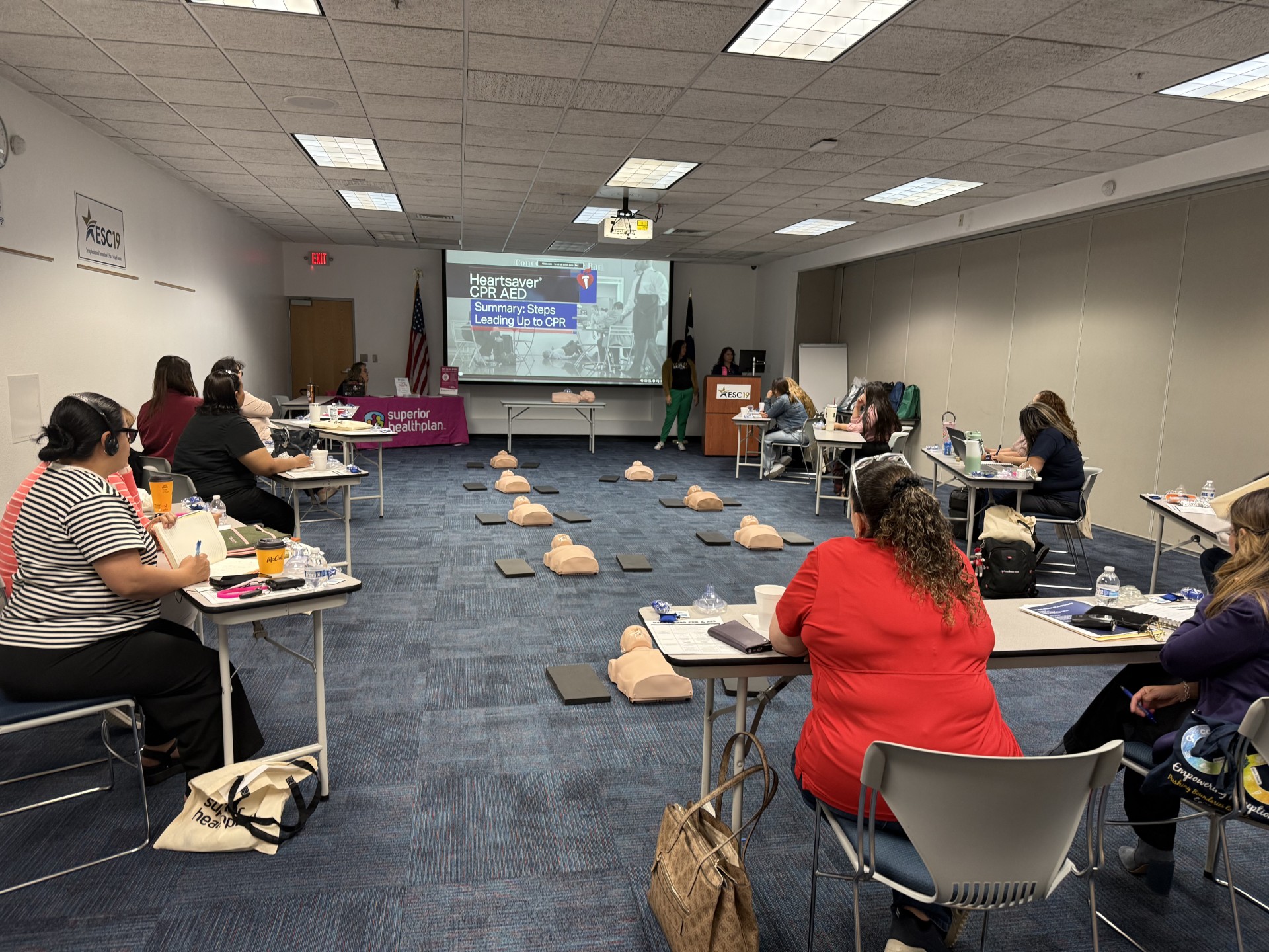 In the foreground, people sit at tables with CPR manikins laid out on the blue carpeted floor, while in the background, a presentation is projected onto a screen.