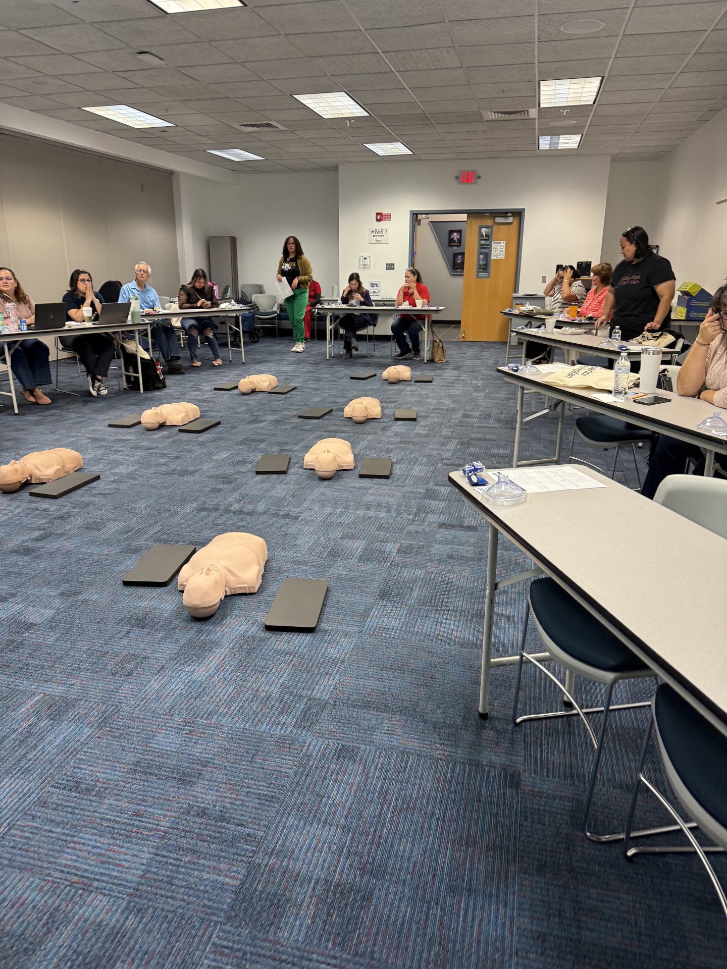 CPR training mannequins are scattered on the floor in the foreground, while people sit at tables in the background, attending a class.