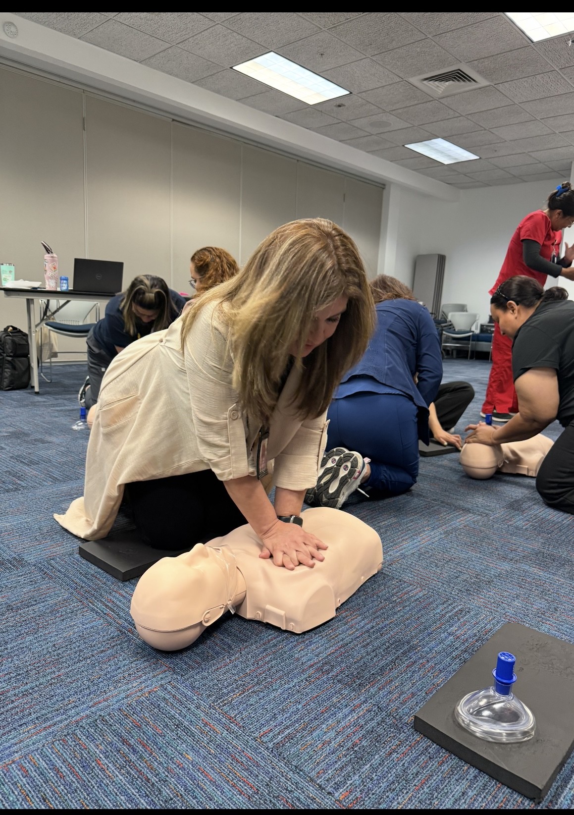 In the foreground, a woman practices CPR on a manikin, while in the background, other participants observe and engage in a training session.