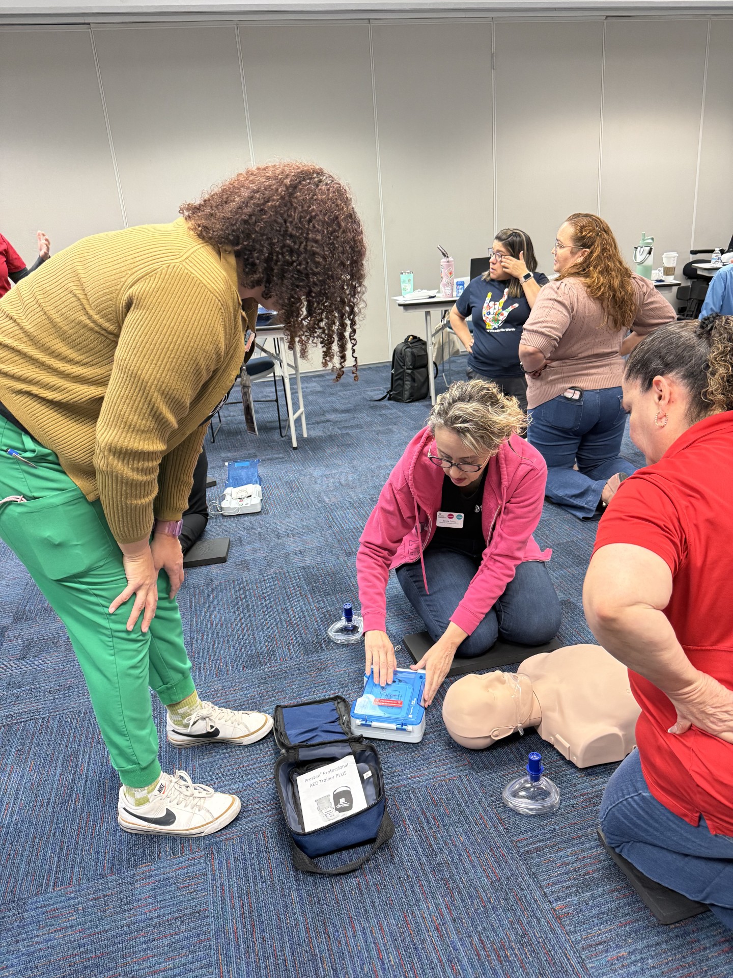 In the foreground, people practice CPR on a mannequin, while in the background, others observe and participate in a training session.