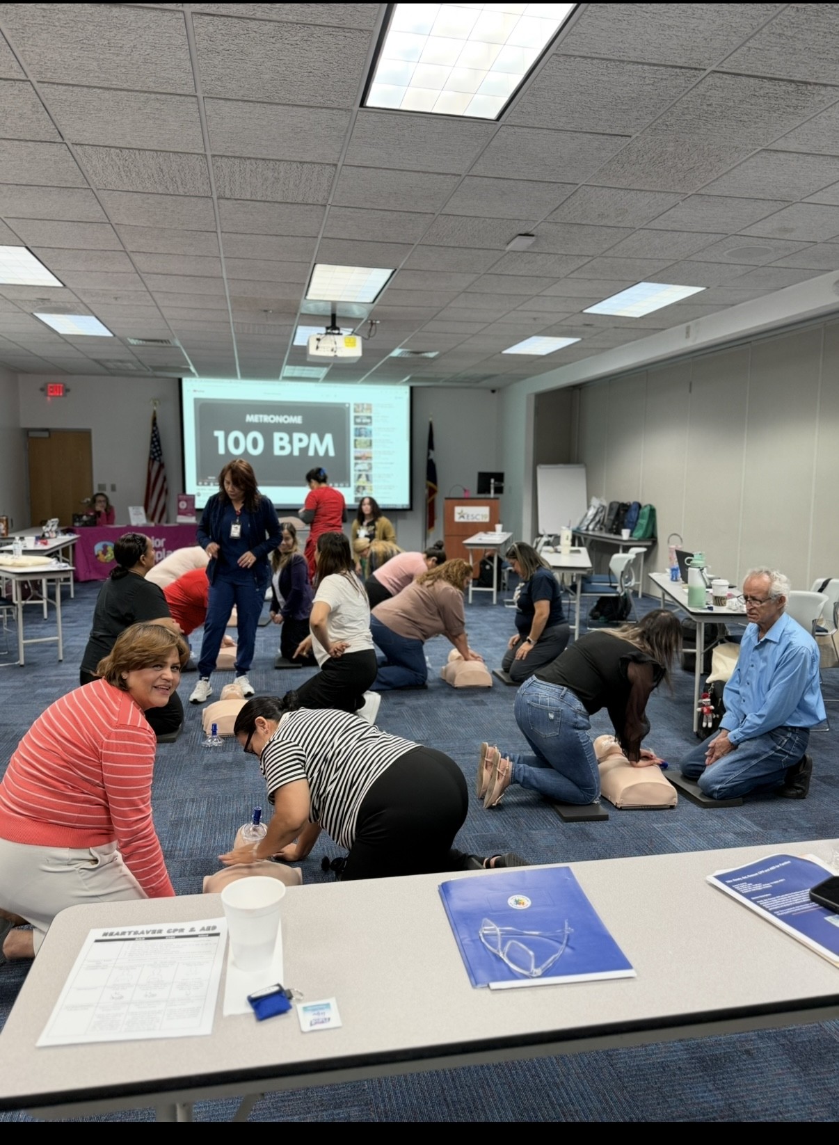 In the foreground, people are practicing CPR on manikins, while in the background, a screen displays "100 BPM" and a projector hangs from the ceiling.