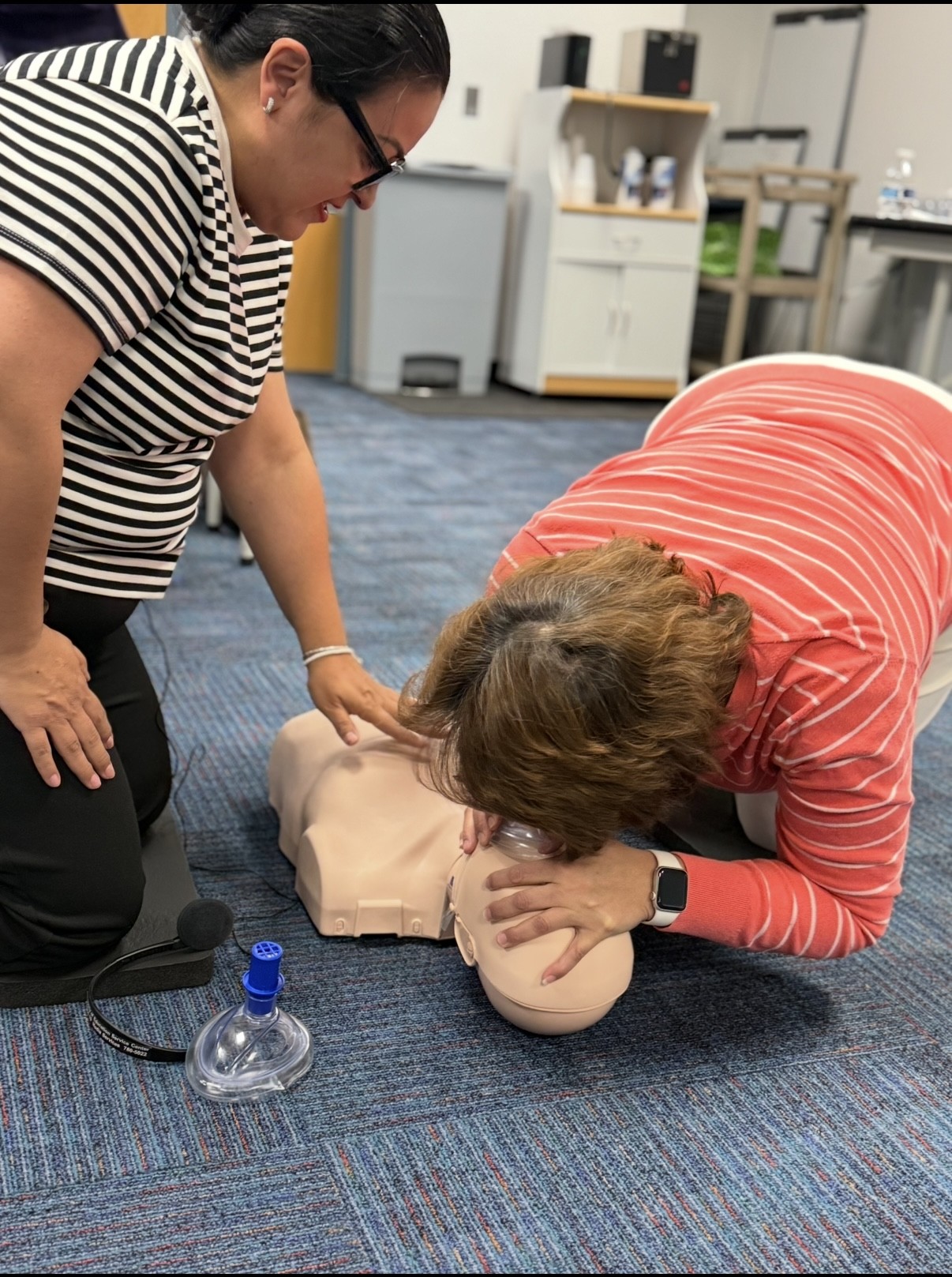 Two women practice CPR on a manikin in the foreground, with a microphone and resuscitation mask nearby, while a blurred background shows a classroom setting with cabinets and chairs.