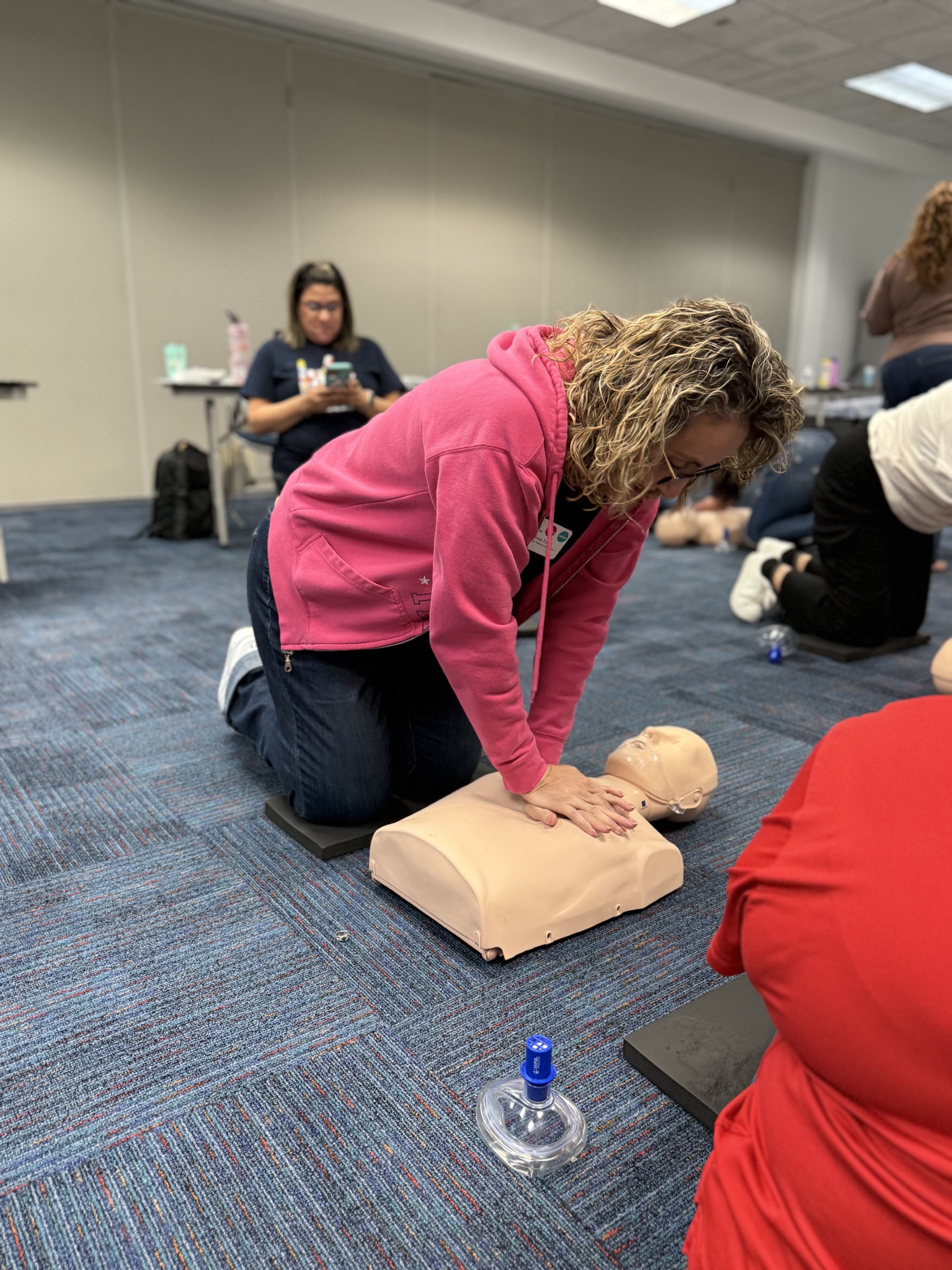 A woman in a pink hoodie practices CPR on a dummy in the foreground, while other participants and instructors are visible in the blurred background of a training room.