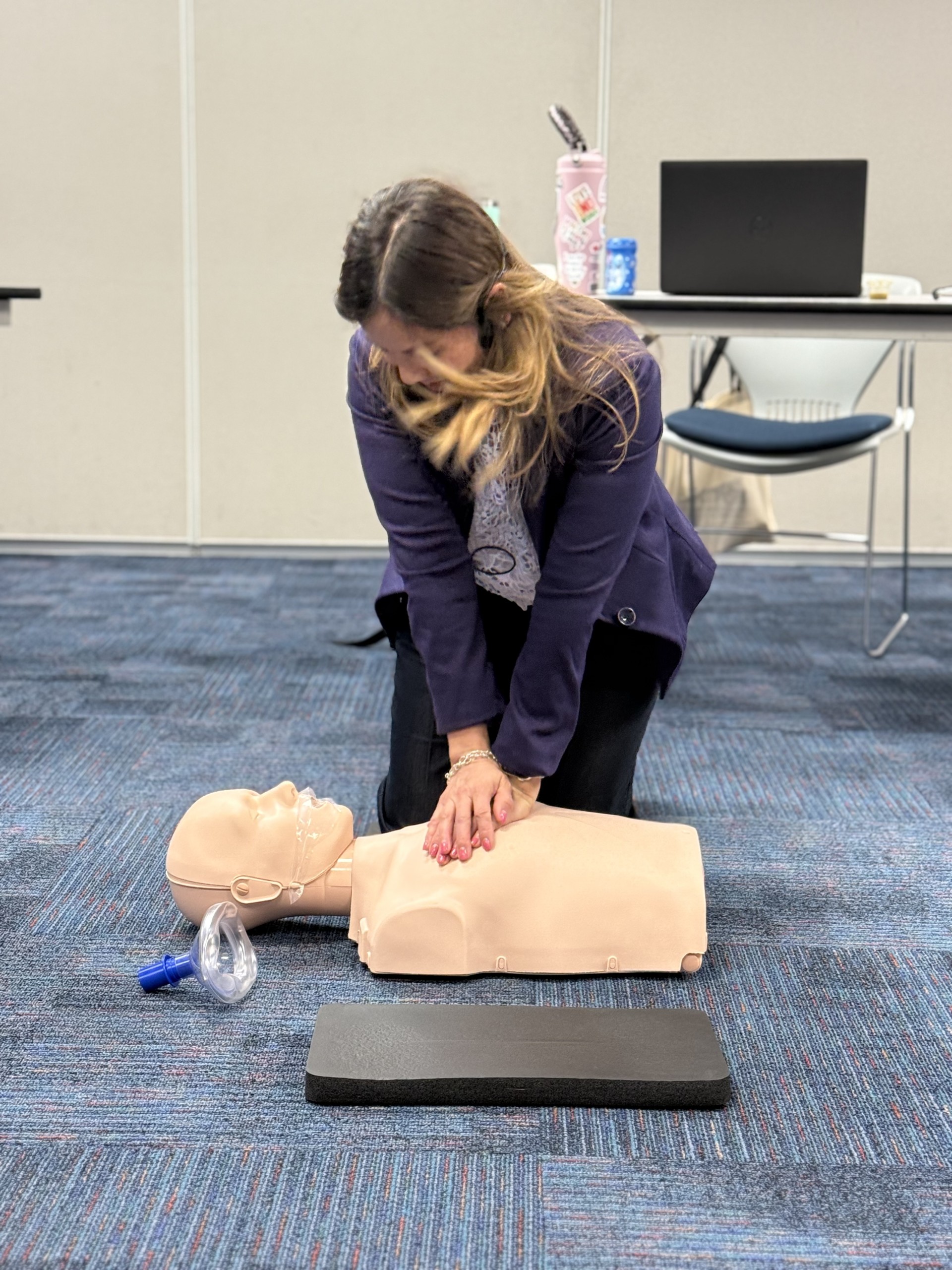 A woman practices CPR on a manikin on a blue carpeted floor, with a table and laptop visible in the background.