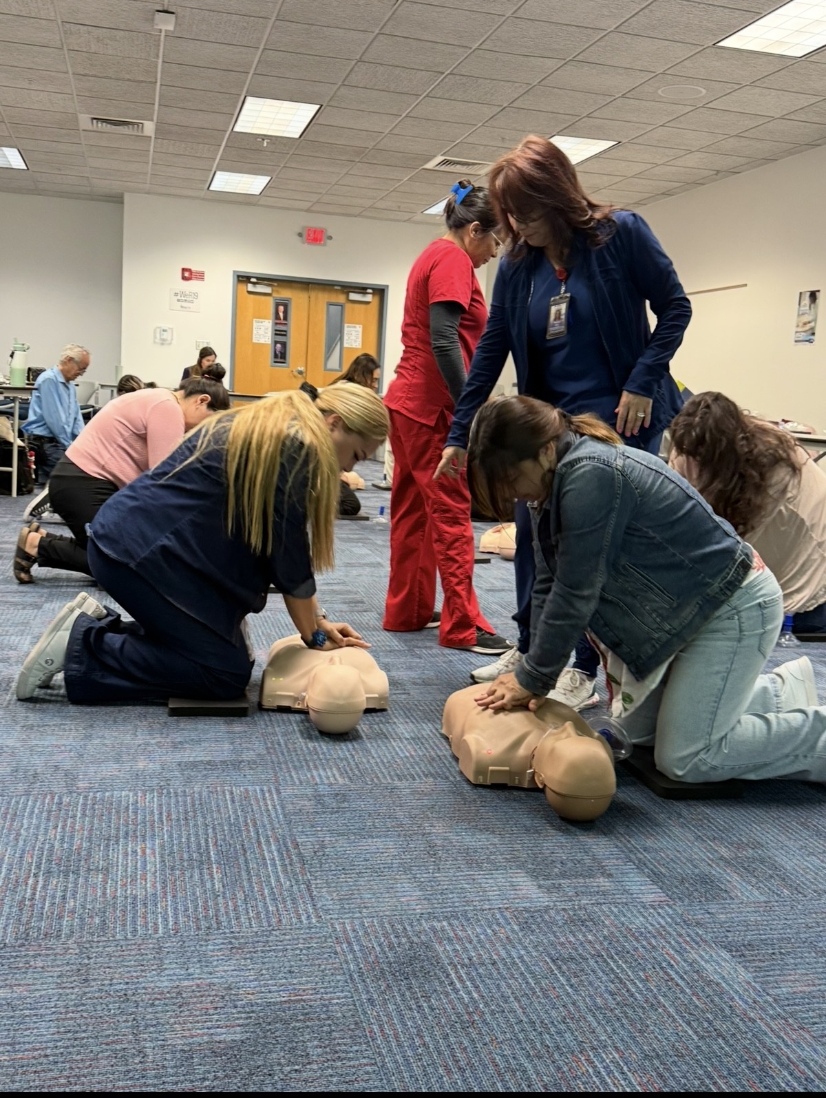 In the foreground, several people practice CPR on mannequins, while in the background, others observe and participate in a classroom setting.