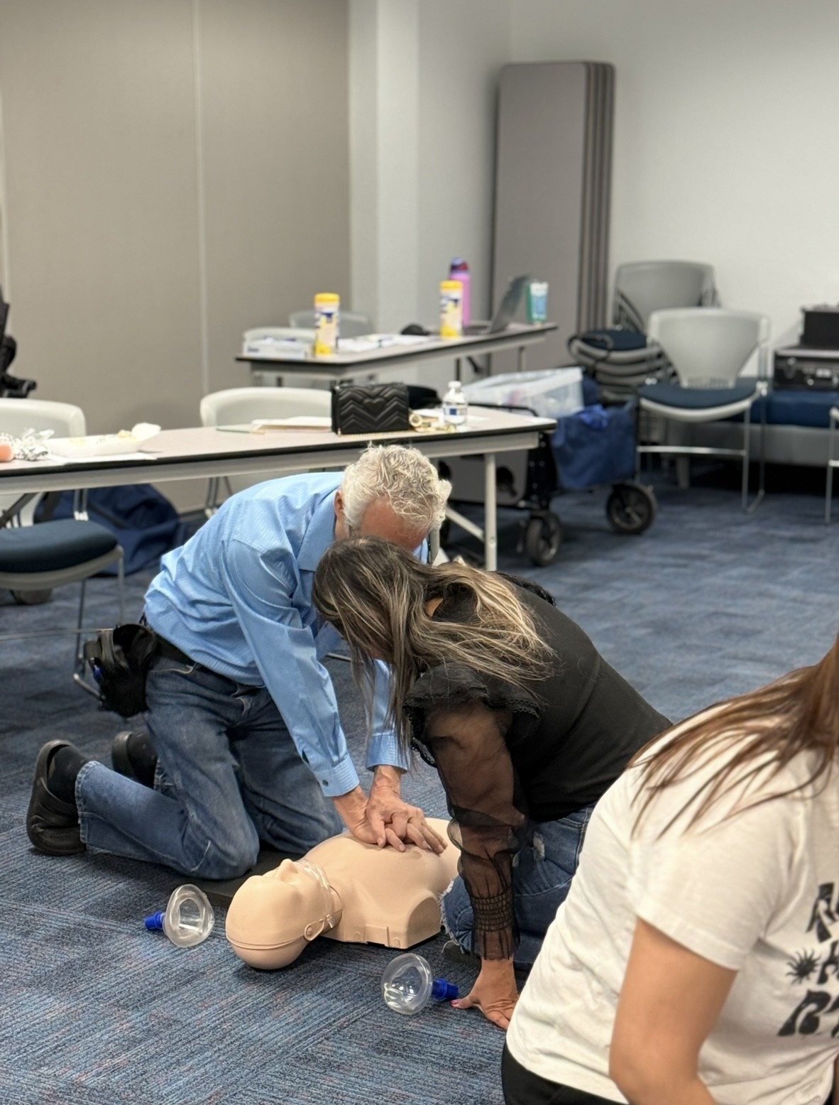 In the foreground, two people practice CPR on a dummy, while the background shows a classroom setting with tables and chairs.