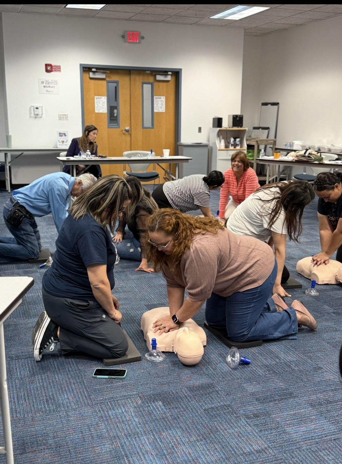 Several people practice CPR on manikins in the foreground, while others observe and a whiteboard is visible in the background.