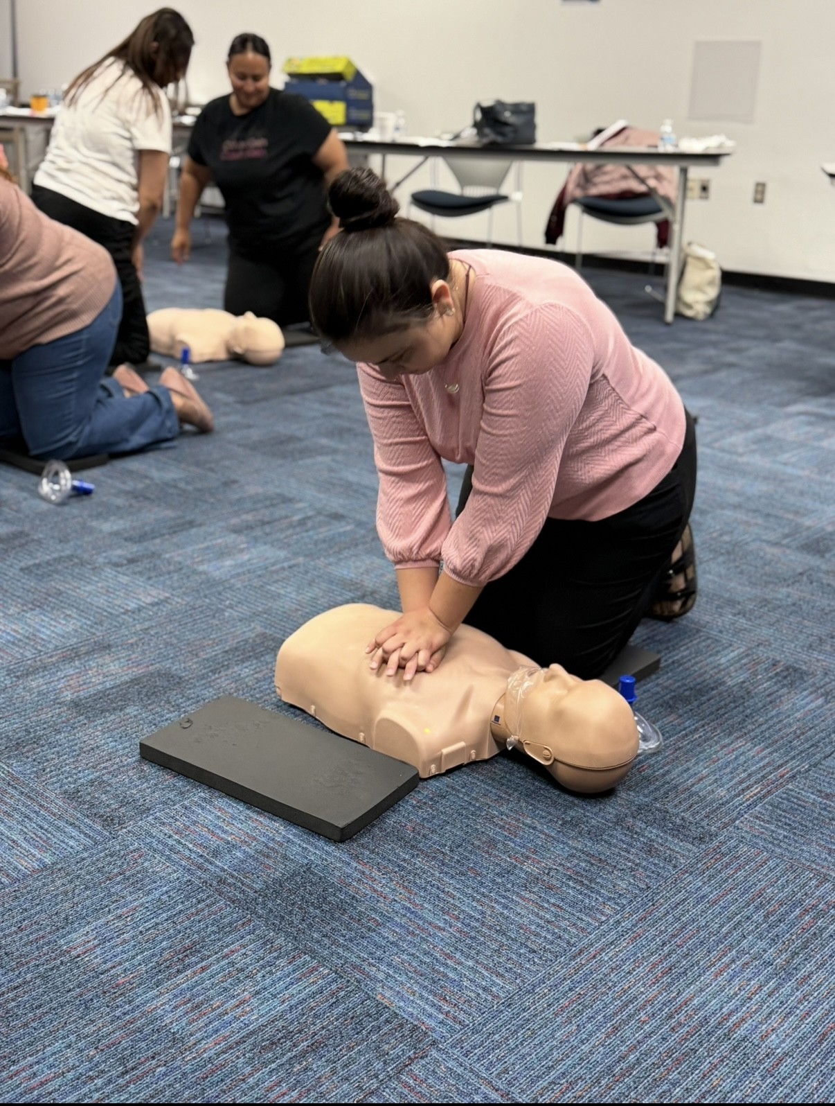 A woman in a pink shirt practices CPR on a manikin in the foreground, while other participants and training equipment are visible in the background.