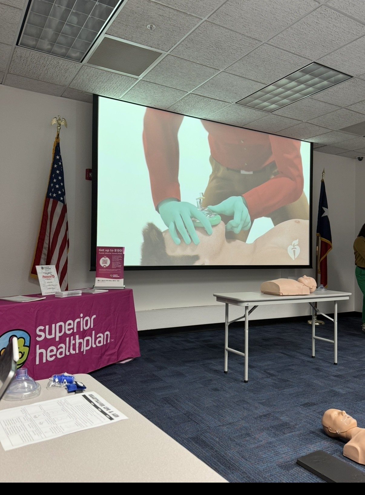 In the foreground, a table displays CPR training materials and mannequins, while the background features a large projection screen showing a CPR demonstration and American flags.