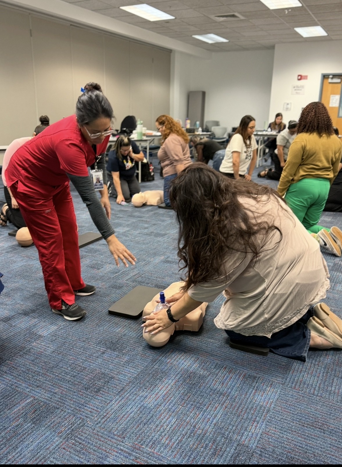 In the foreground, a woman in red scrubs instructs another woman on performing CPR on a dummy, while in the background, other participants practice on similar dummies in a classroom setting.
