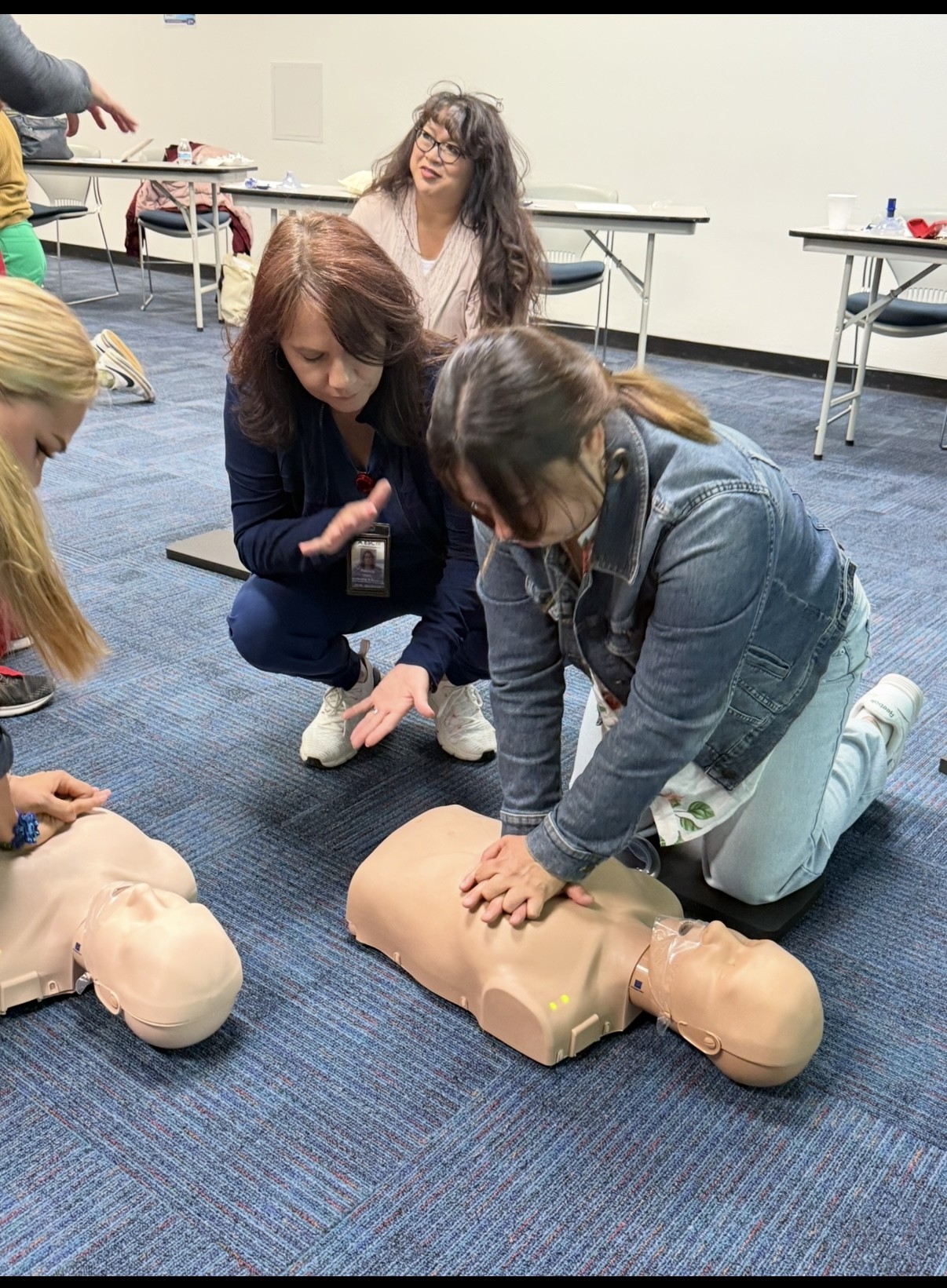 In the foreground, several women practice CPR on training mannequins, while in the background, other participants observe and tables are set up in a classroom.