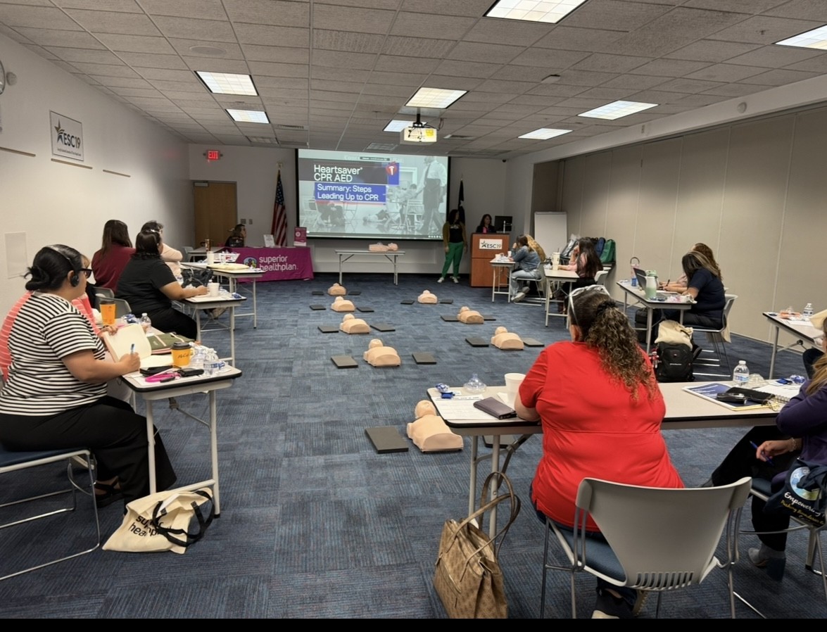 People in the foreground are seated at tables with CPR manikins laid out on the floor in the background, where a presentation is being projected onto a screen.