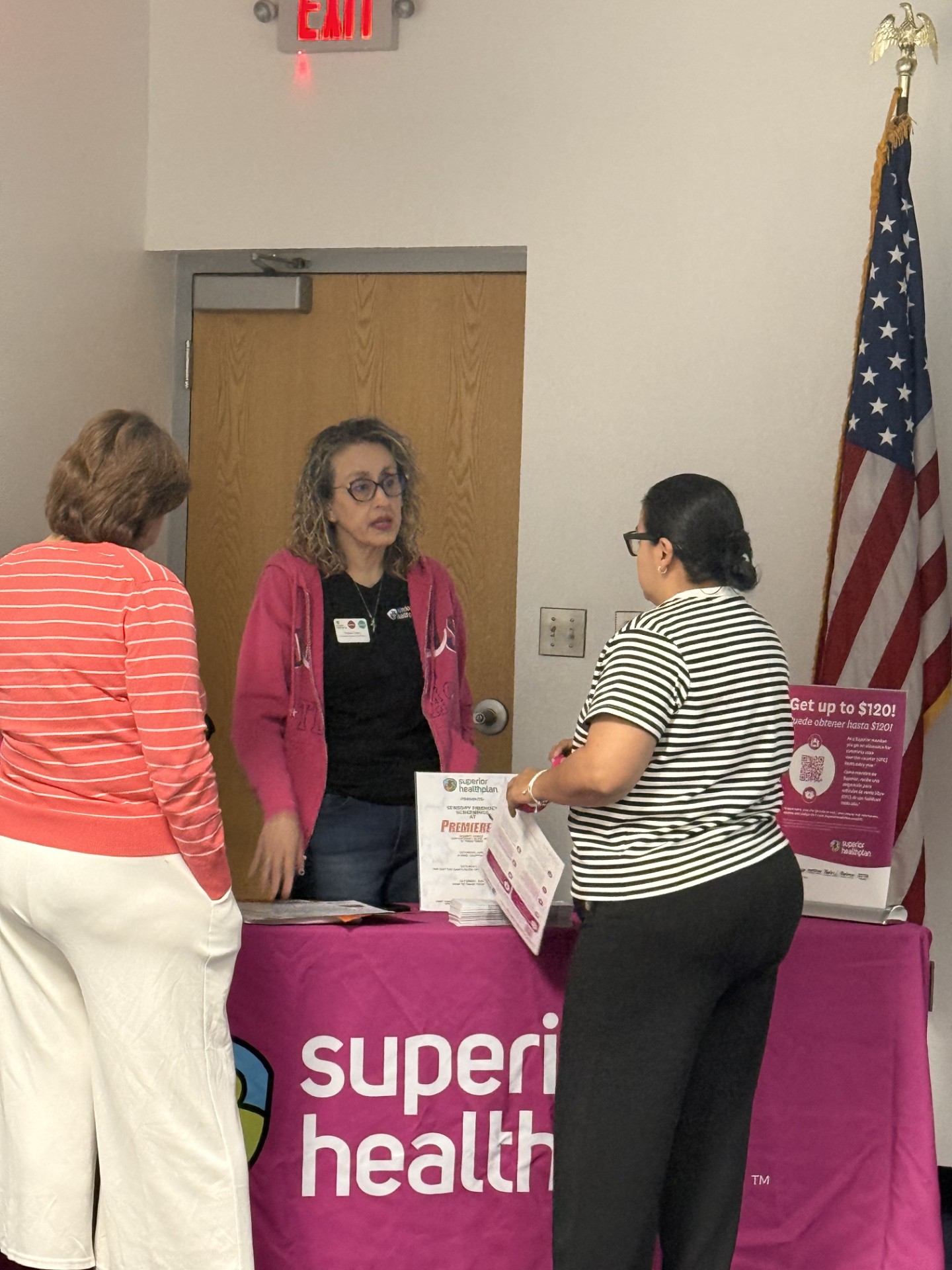 Two women stand at a table with a "superior health" banner, while a third woman in a pink jacket speaks to them, with an American flag and an exit sign in the background.