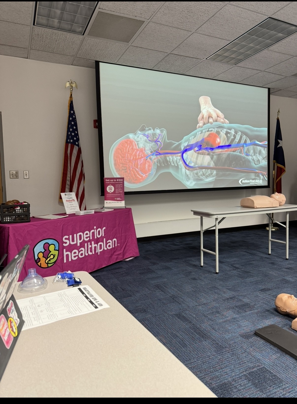 In the foreground, a table displays training materials and CPR mannequins, while the background features a large screen showing an anatomical diagram and American flags.