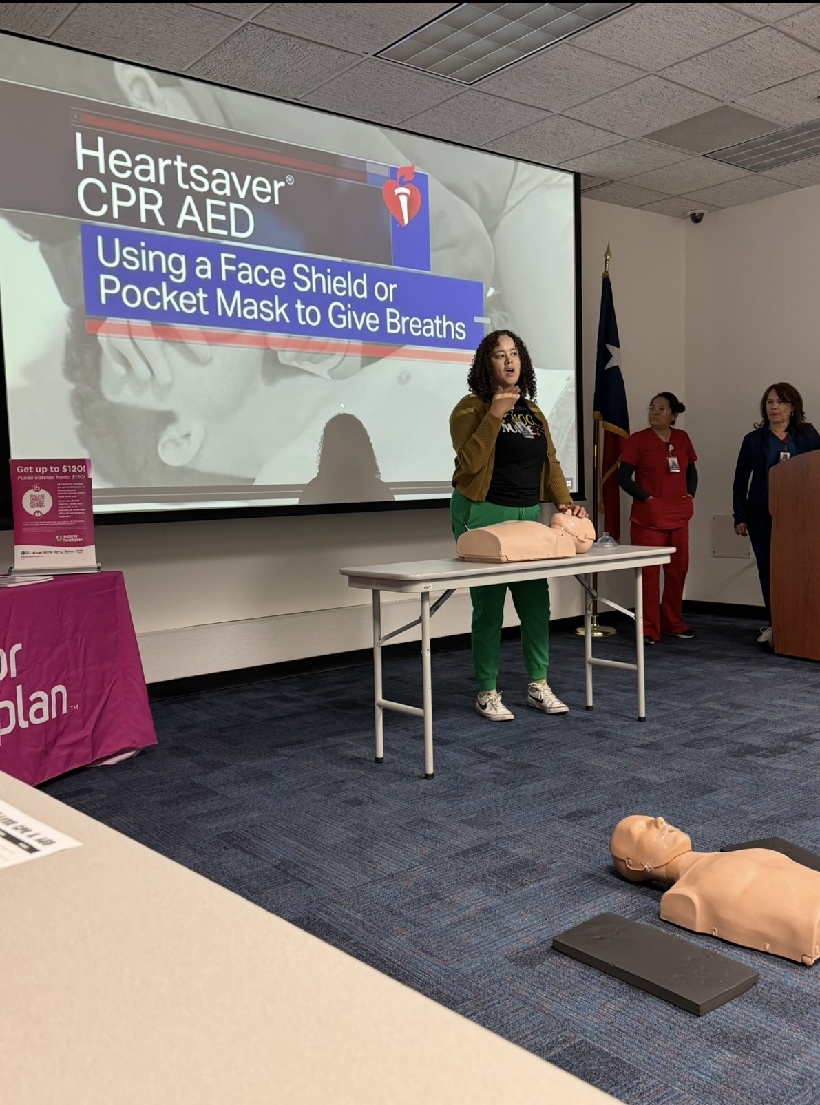 A woman stands in front of a projection screen displaying CPR instructions, with CPR mannequins on the floor in the foreground and other people observing in the background.