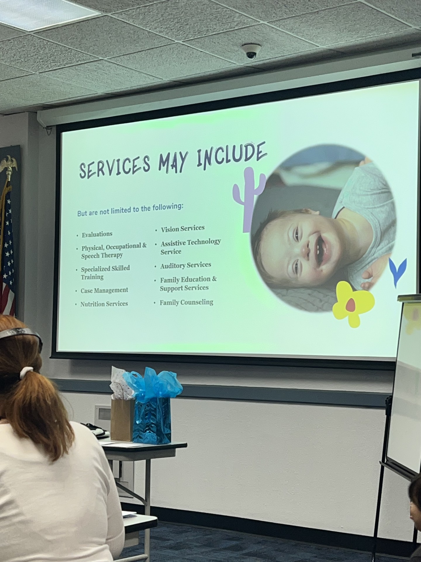 A projector screen displays a list of services with a smiling child in the background, while a woman with her back to the camera sits in the foreground.