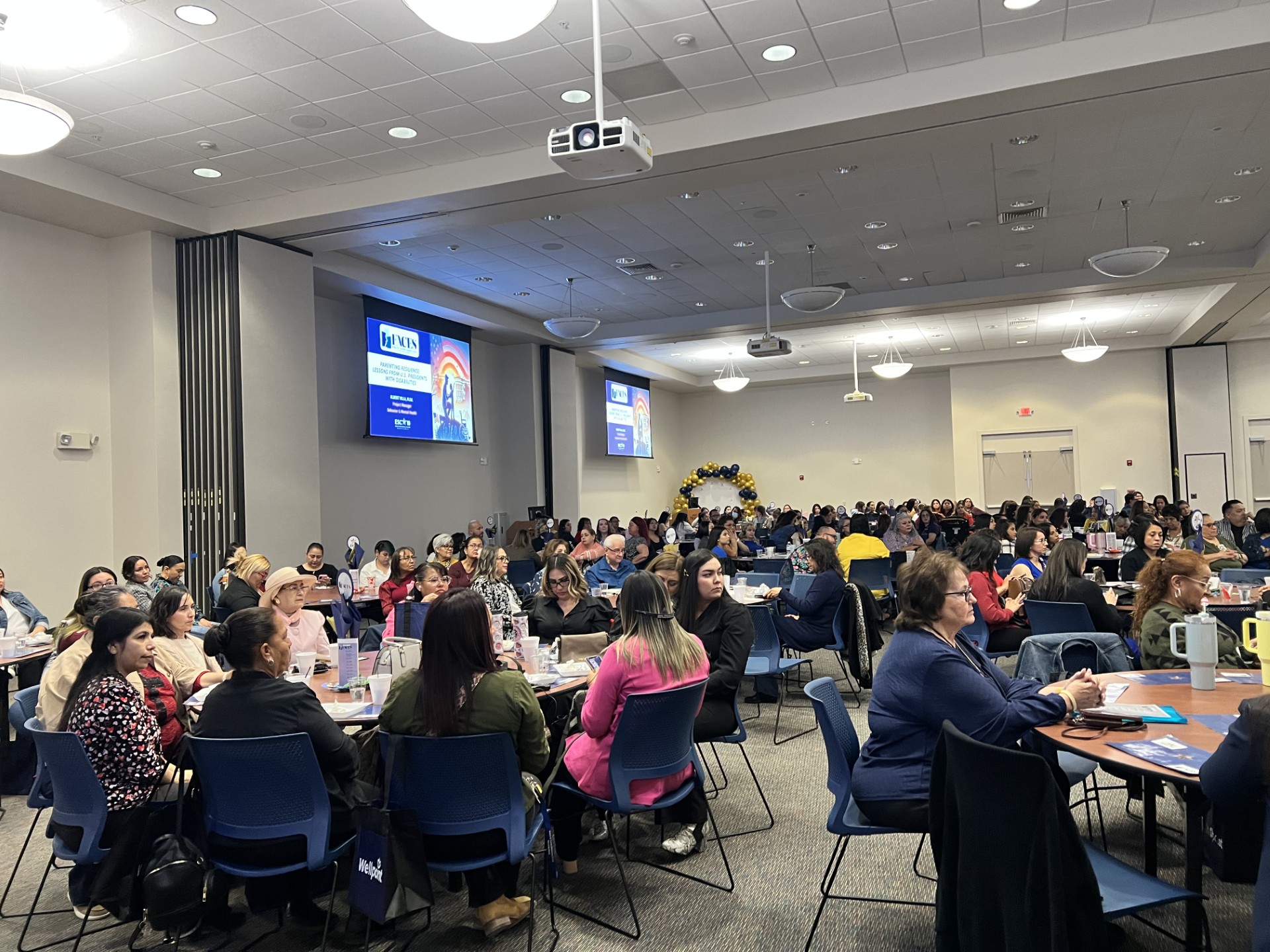 A large audience is seated at tables in a brightly lit conference room, with screens displaying presentations in the background.