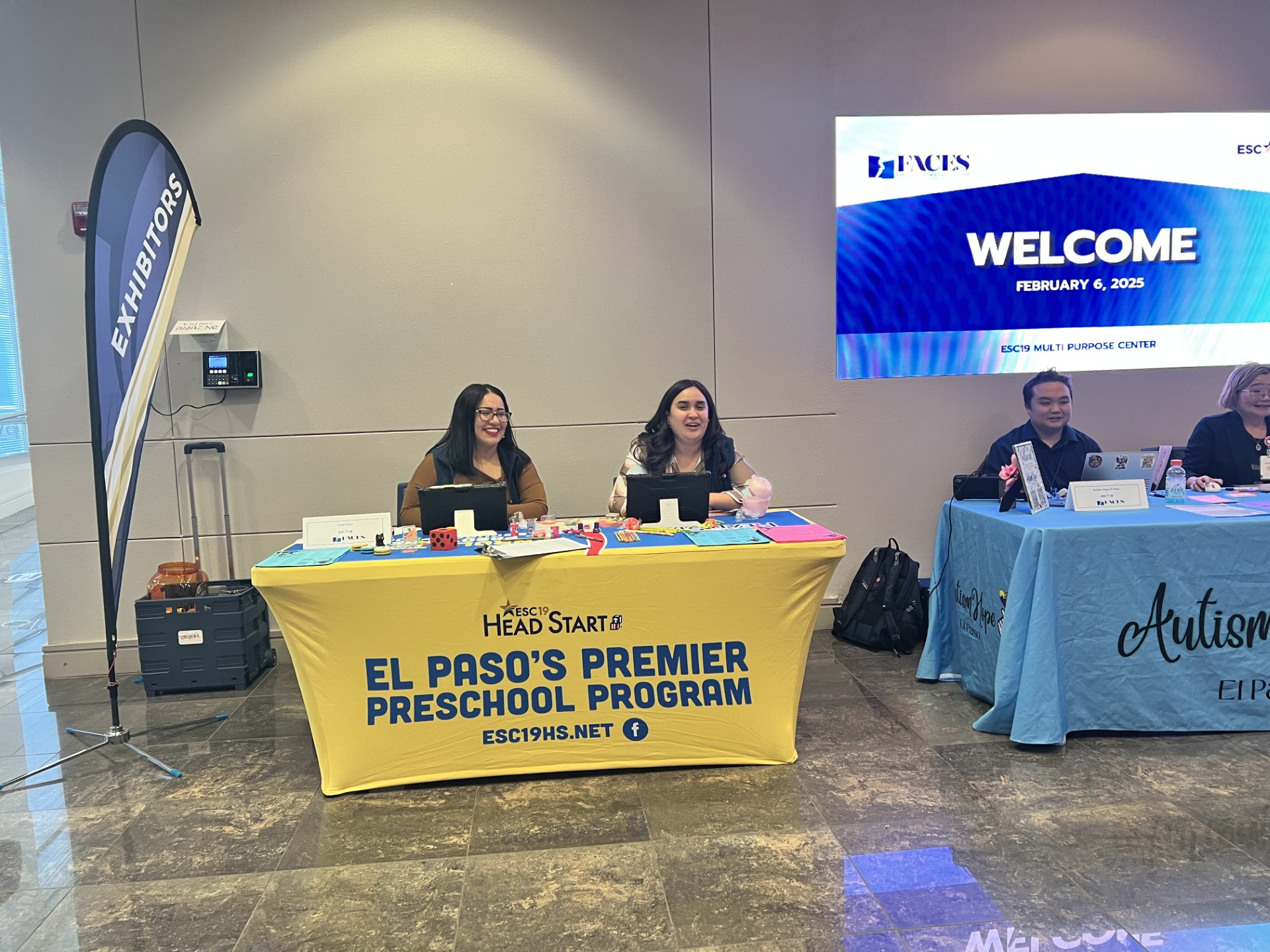 Two women smile behind a yellow table with "El Paso's Premier Preschool Program" printed on it, while a large screen in the background displays "WELCOME FEBRUARY 4, 2025."