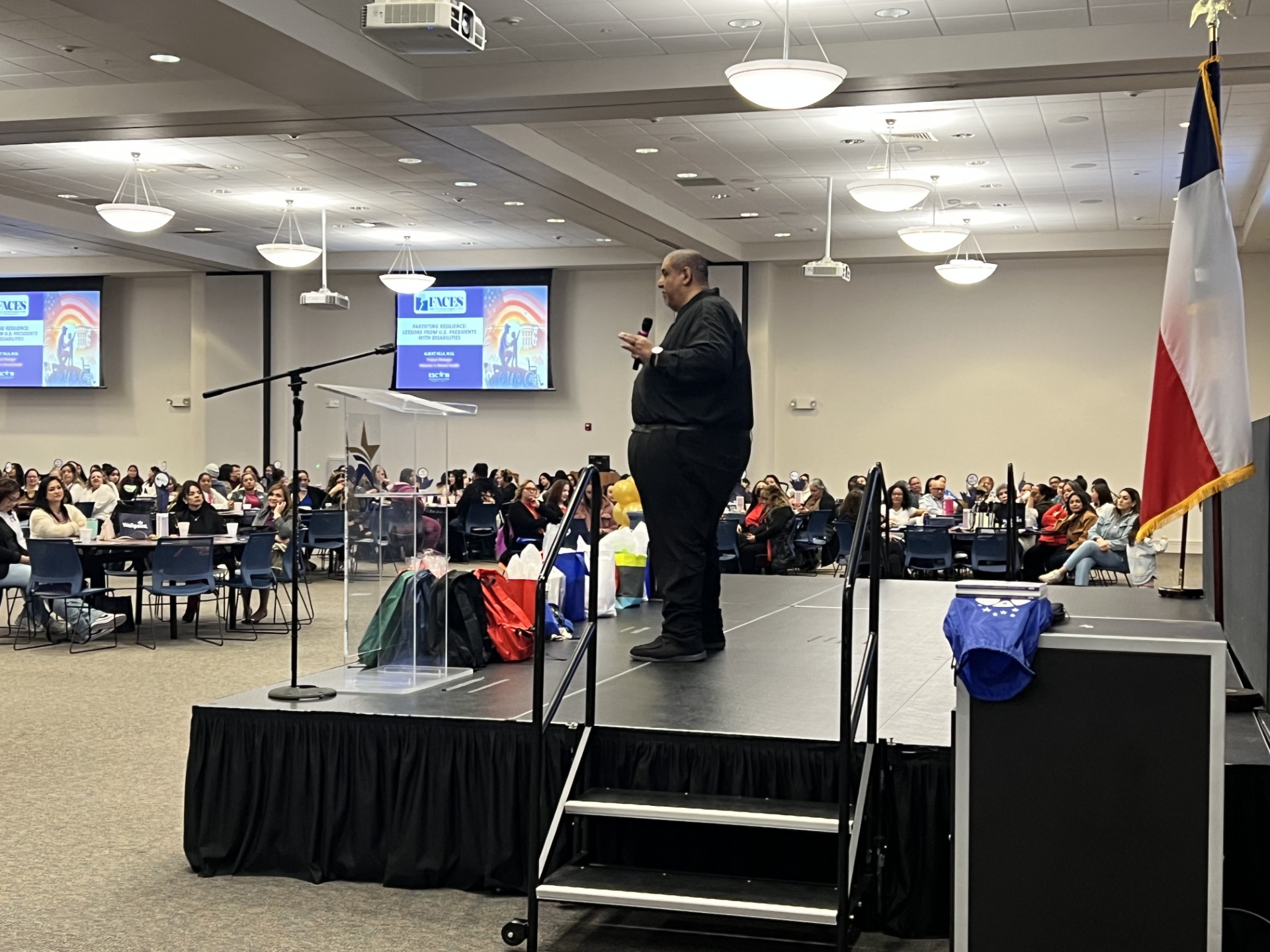 A man in a black shirt and pants speaks from a stage in the foreground, while a large audience sits at tables in the background of a convention hall.