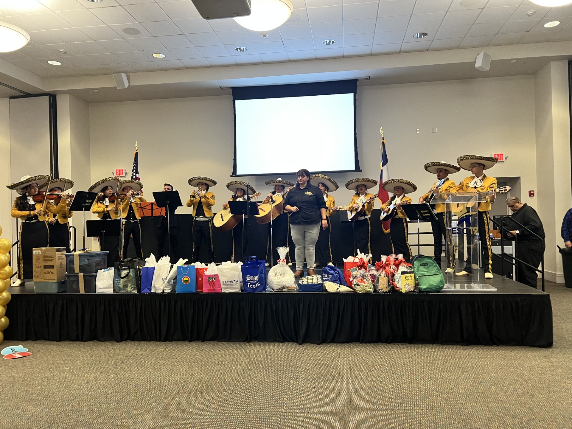 A mariachi band performs on a stage adorned with gifts, while a blank screen hangs in the background.