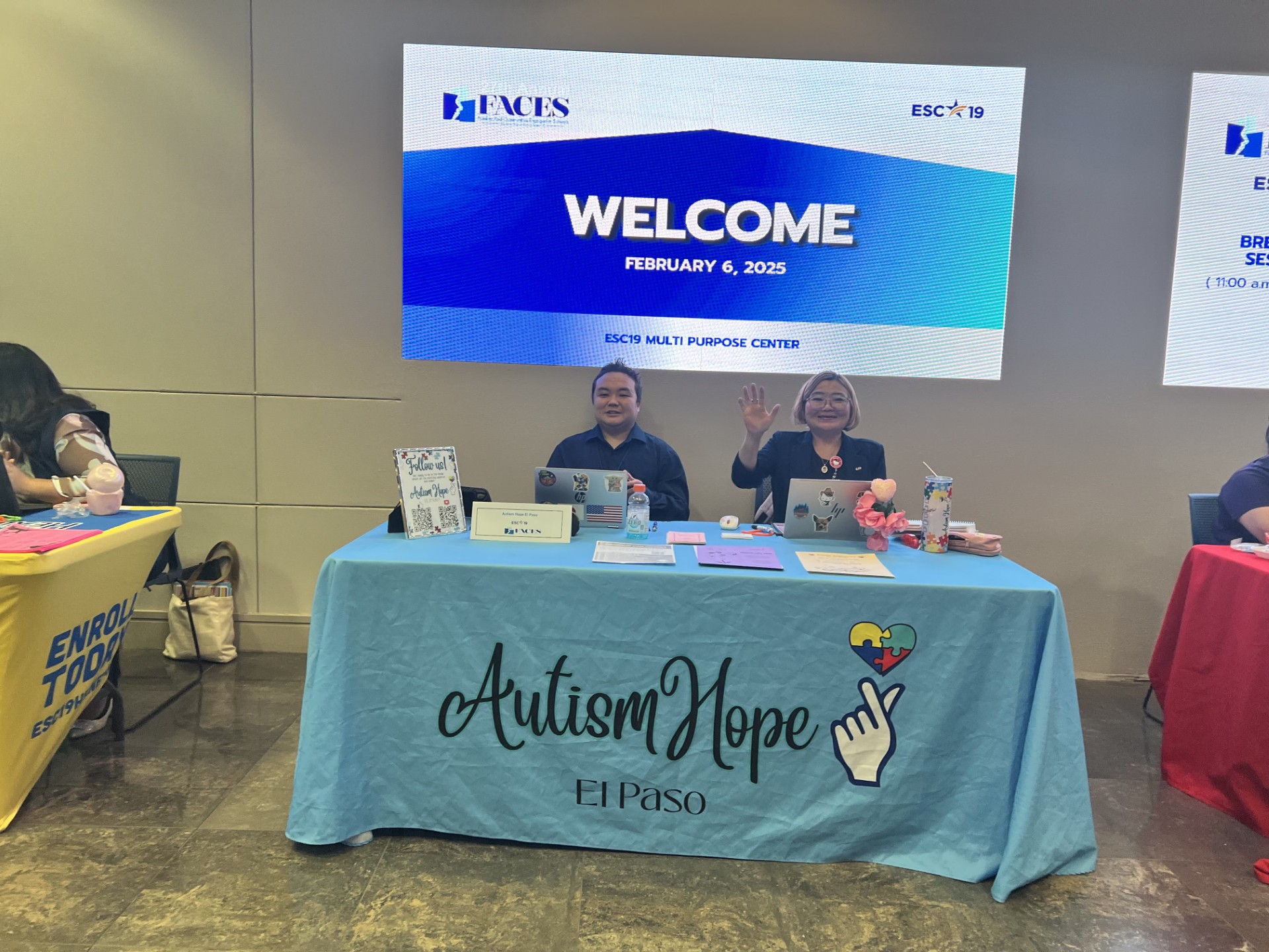 Two women wave from behind a table with "Autism Hope El Paso" written on it, while a large screen in the background displays "WELCOME FEBRUARY 6, 2025".