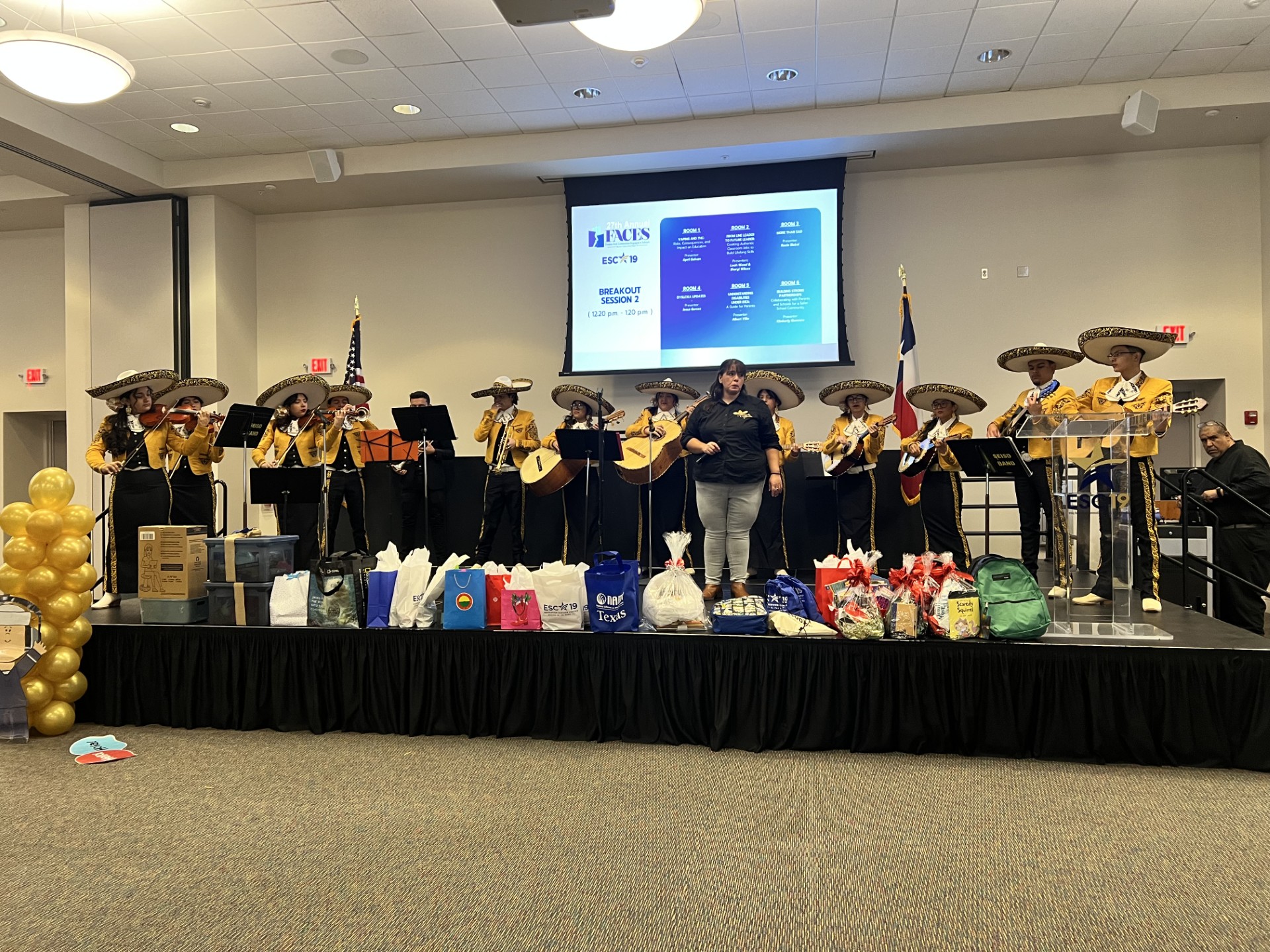 A mariachi band performs on a stage adorned with gifts and balloons, with a presentation screen displaying information in the background.