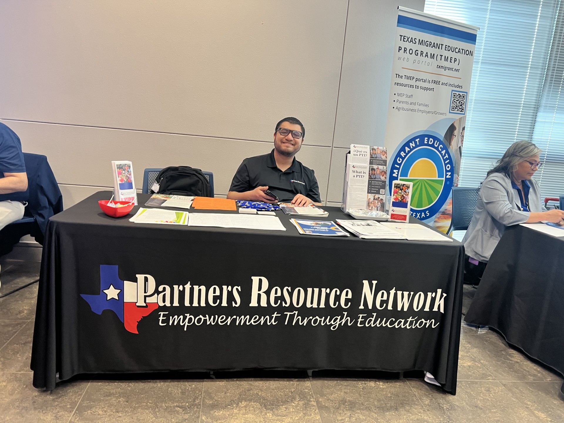 A man smiles from behind a table with a "Partners Resource Network" tablecloth, while a banner for the "Texas Migrant Education Program" stands in the background.