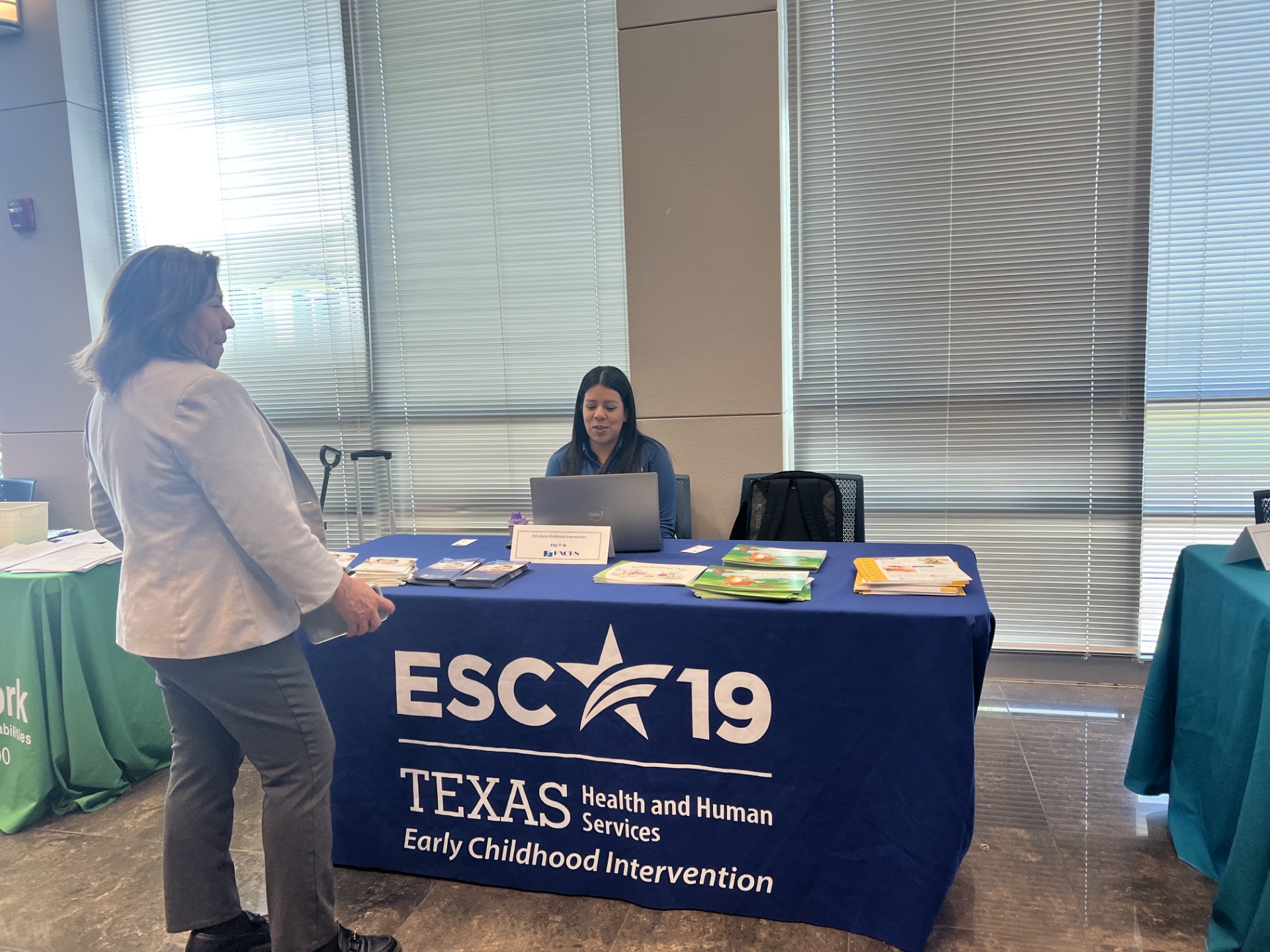 A woman in a light jacket stands at a table with a blue tablecloth displaying "ESC 19 Texas Health and Human Services Early Childhood Intervention" in the foreground, while a woman sits behind the table with a laptop in the background.