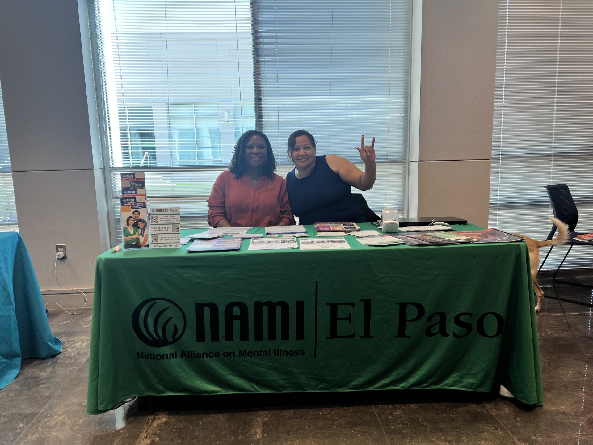 Two women stand behind a green table with "NAMI El Paso" printed on it, while blinds cover large windows in the background.