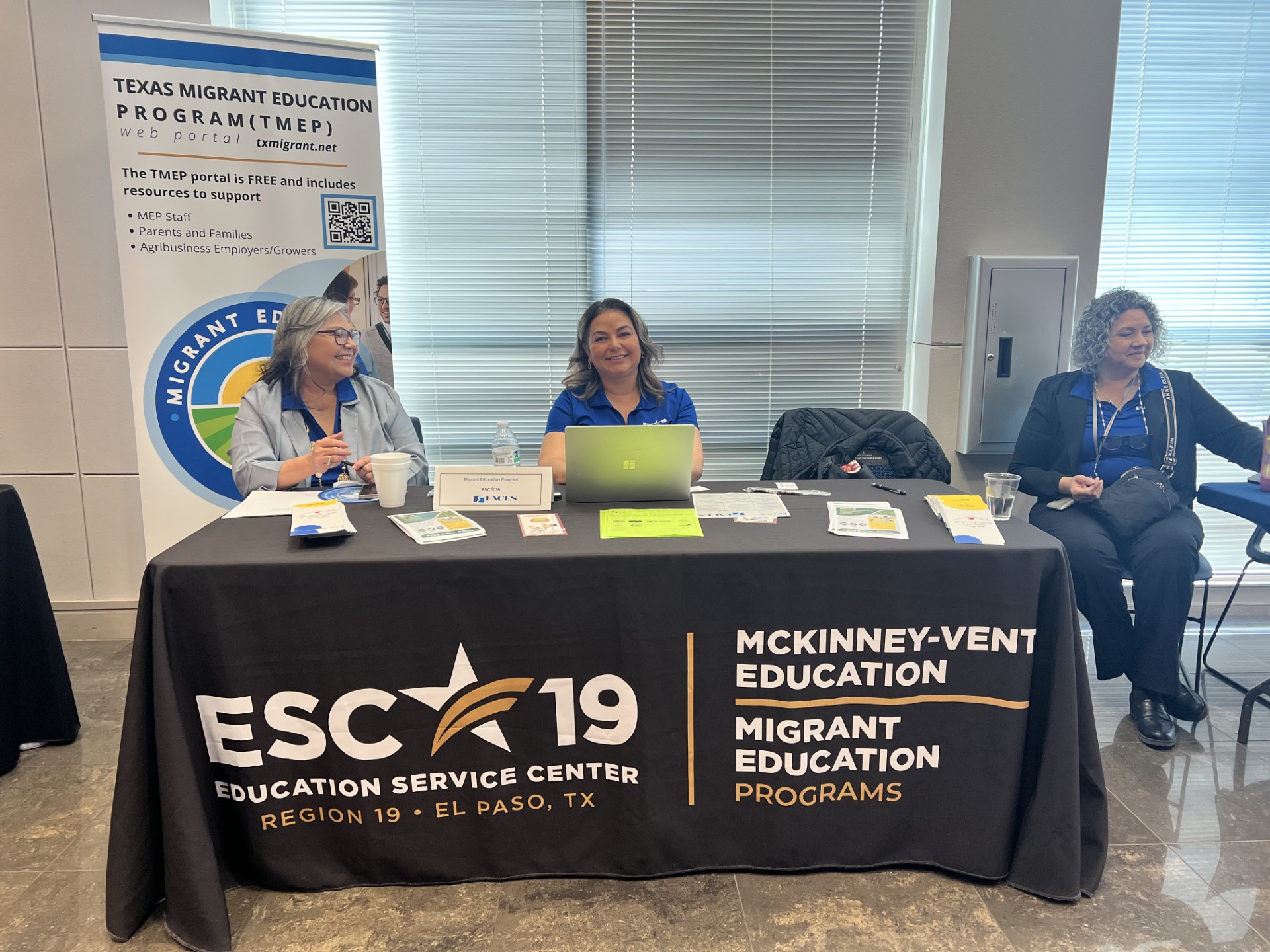 Three women staff a table with promotional materials for the Texas Migrant Education Program in front of a banner.