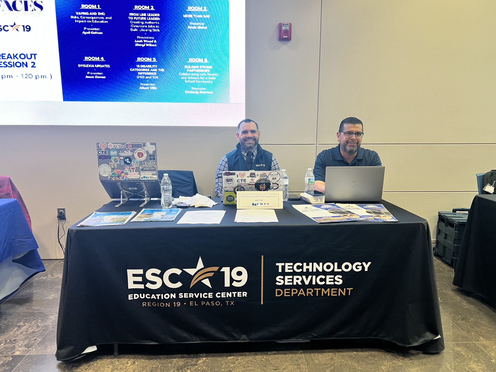 Two men stand behind a table with the ESC 19 logo, while a large screen displaying event information is visible in the background.