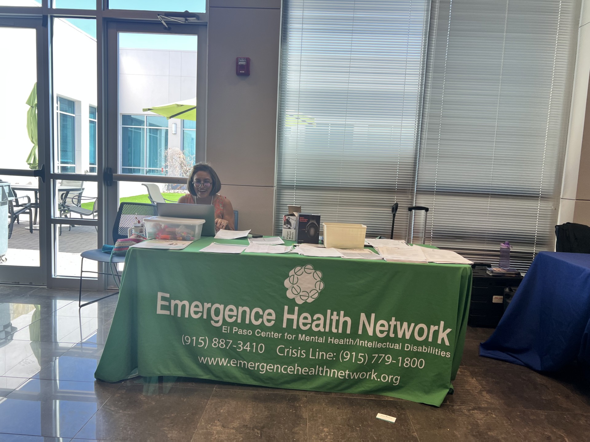 A woman sits behind a table with a green tablecloth displaying "Emergence Health Network" in the foreground, while large windows and outdoor seating are visible in the background.